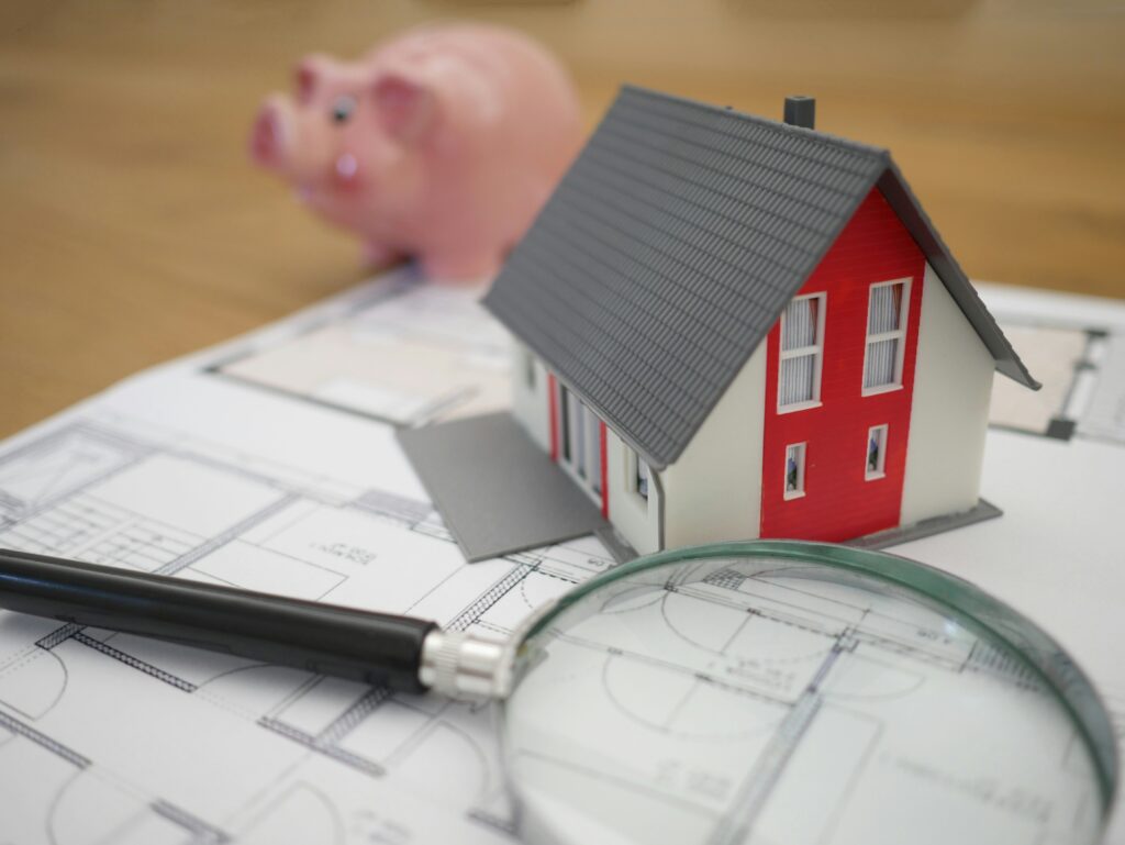 Small model house sits on top of blueprints with piggy bank in the background. 